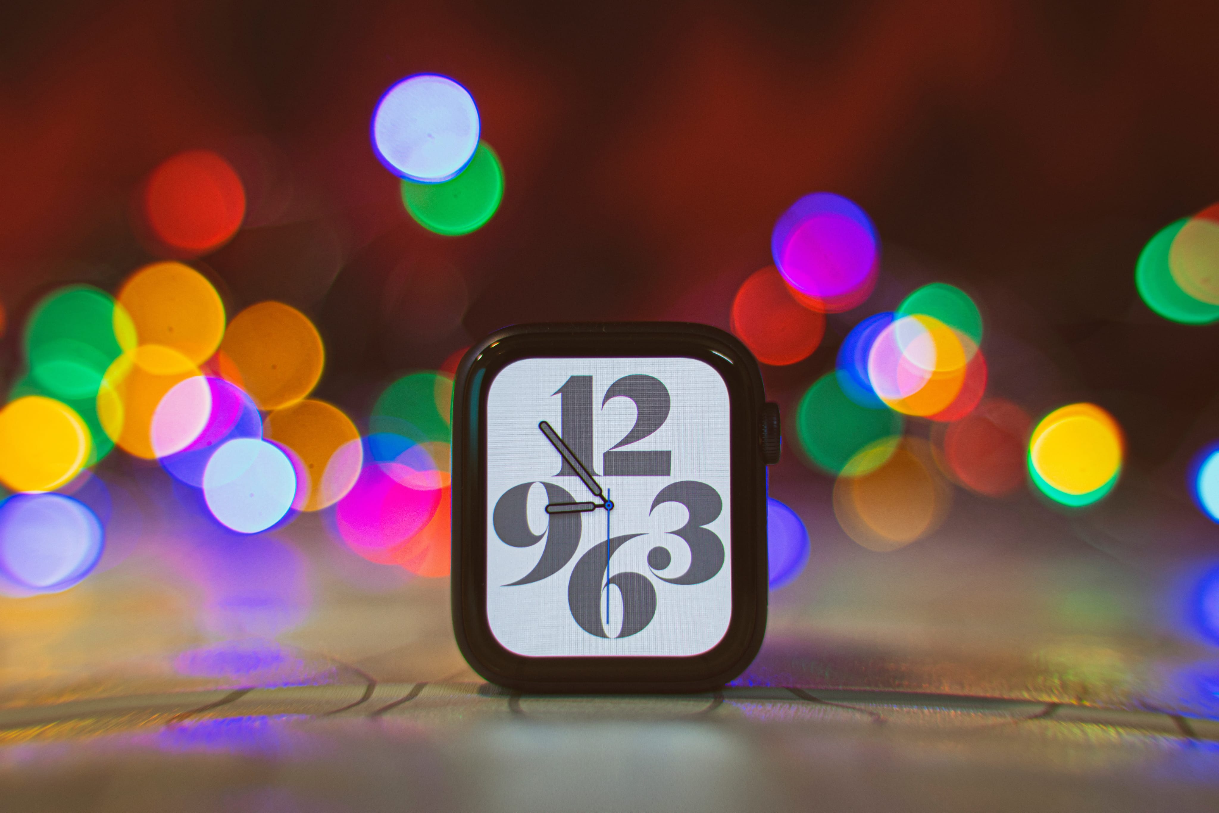 A photograph showing an Apple Watch Series 6 case sitting upright on a table, with a colorful lights in the background blurred with a depth-of-field effect
