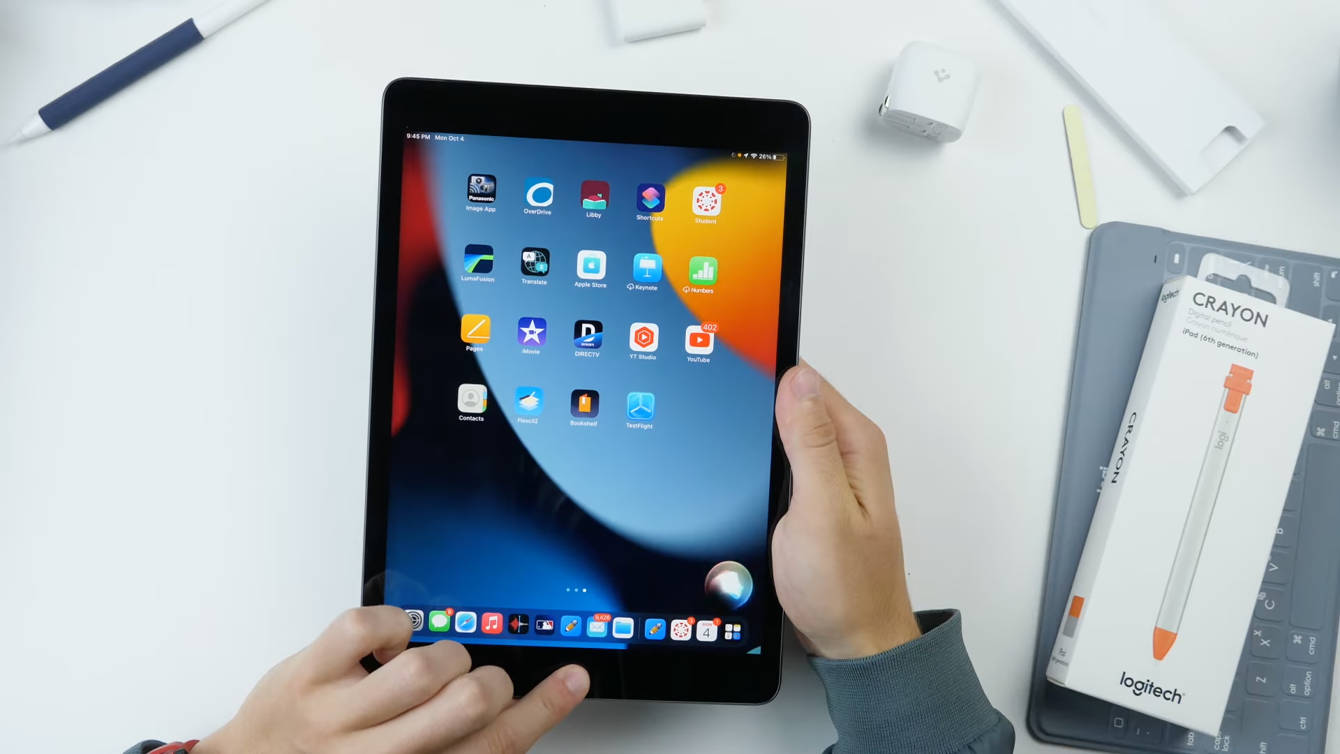 Une vue de haut en bas d'un bureau blanc avec les mains de votre homme tenant l'iPad de neuvième génération d'Apple dans les mains avec l'écran d'accueil par défaut affiché à l'écran