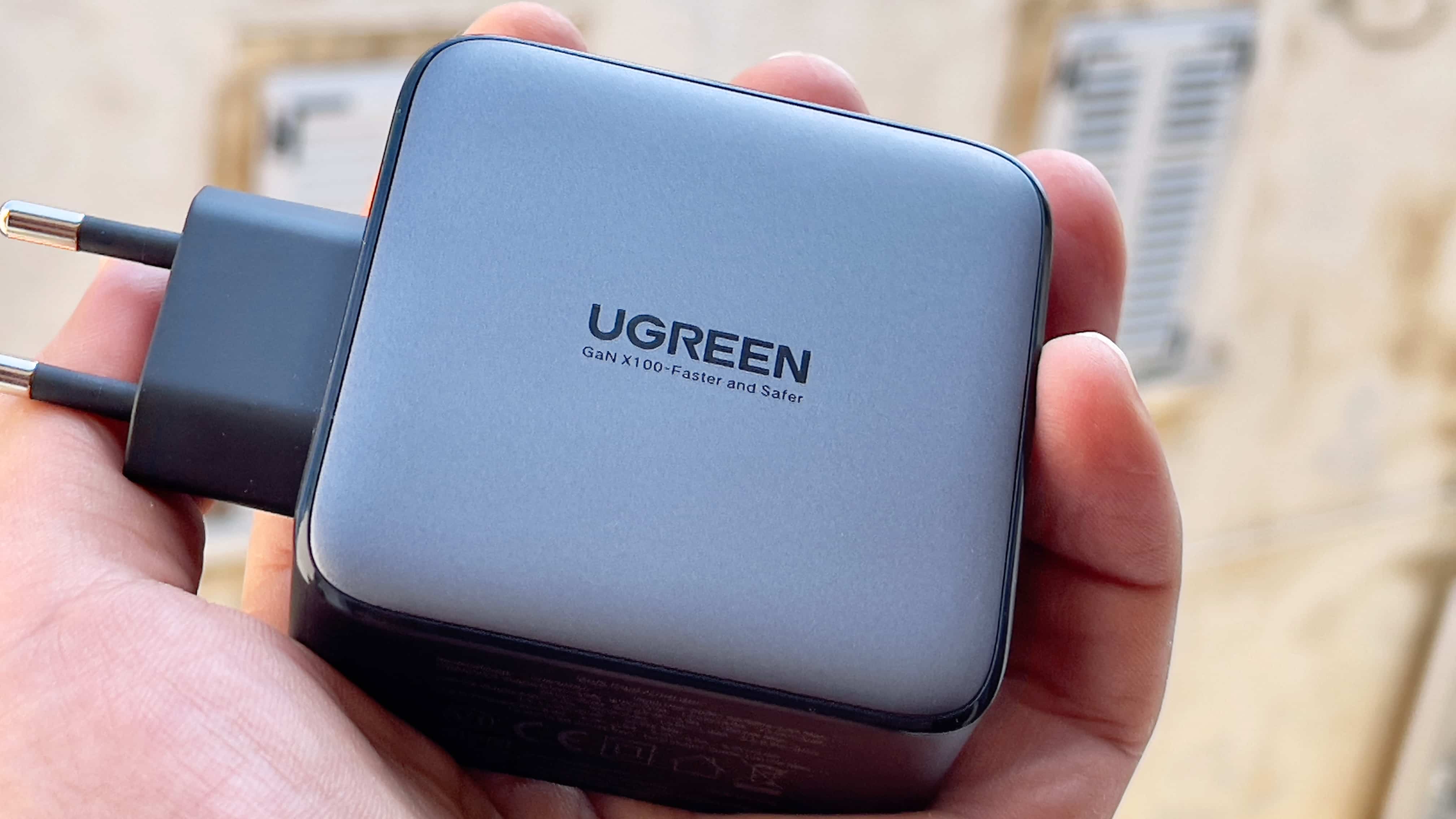 Une main masculine tenant l'adaptateur secteur Nexus de 100 watts d'Ugreen, montrant un logo Ugreen avec le slogan "GaN X100-plus rapide et plus sûr" imprimé au dos du boîtier