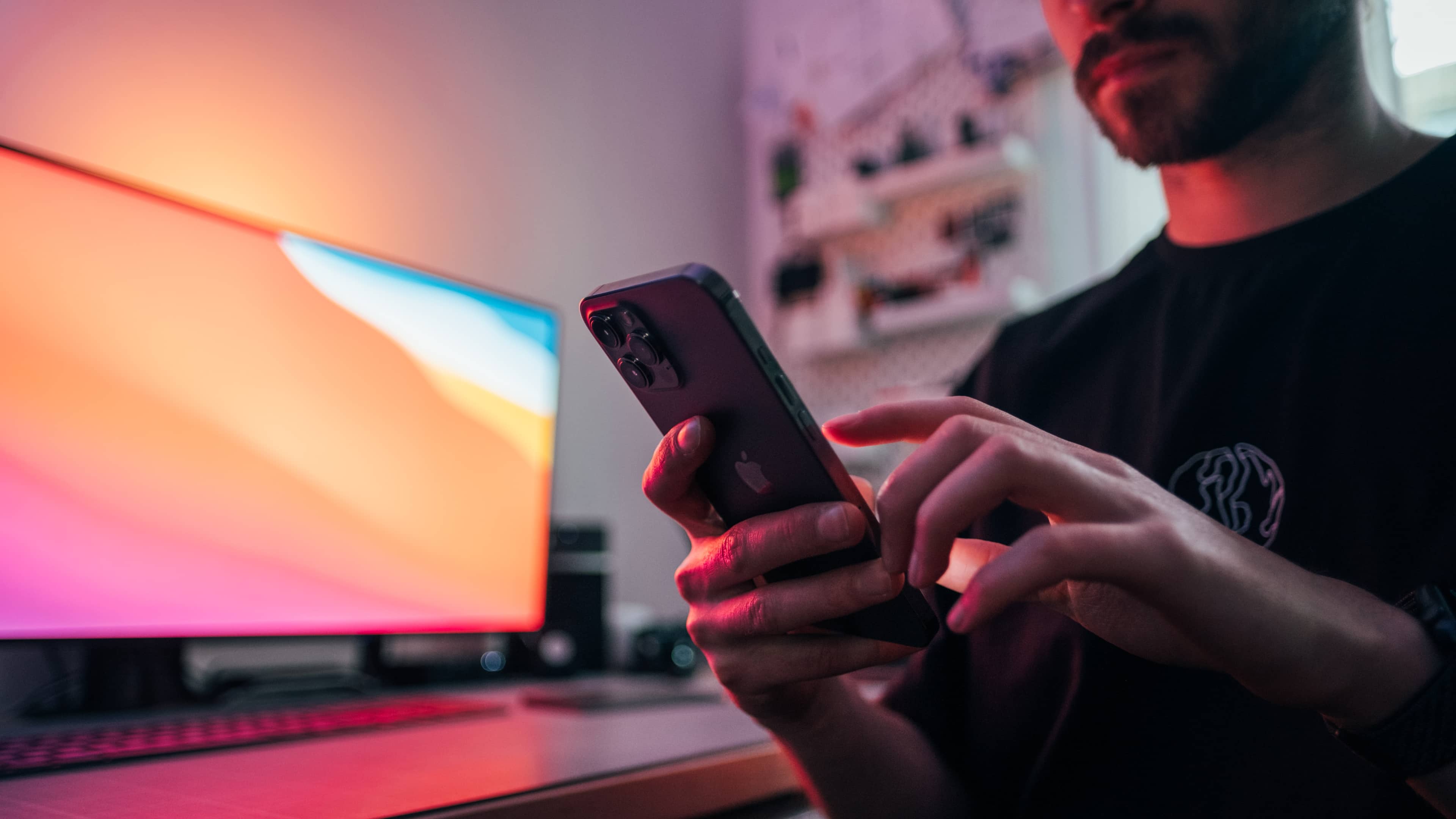 Jeune homme tenant un iPhone 12 Pro Max devant son bureau.  En arrière-plan, il y a un moniteur utilisant le fond d'écran Big Sur et le clavier et le trackpad gris sidéral d'Apple.