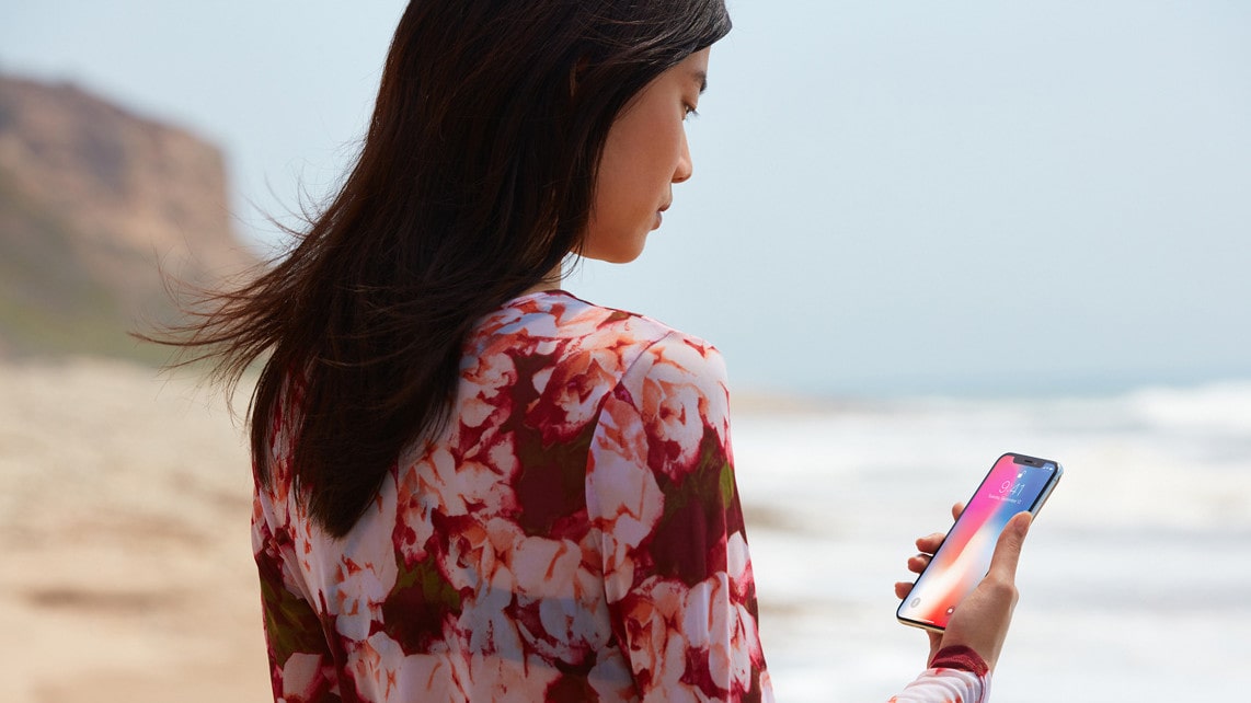 A woman on a beach looks at an iPhone X and unlocks it using Face ID.